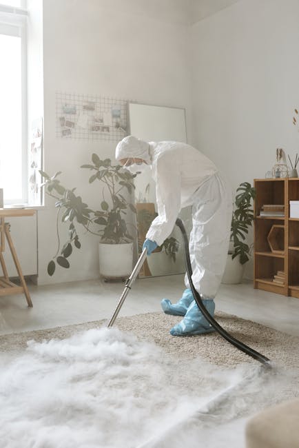 A professional cleaner dressed in full protective gear, including a white suit, gloves, and boots, is using a vacuum extraction machine on a beige carpet in a well-lit, modern living room. The room features white walls, a large window allowing natural light, and various indoor plants in white pots for decor. There is a wooden sideboard with decorative items and a small wooden table near the window. The scene depicts thorough surface cleaning and deep cleaning processes essential for maintaining hygiene, as performed by Hounslow Carpet Cleaning, emphasizing effective carpet sanitisation in a domestic setting.