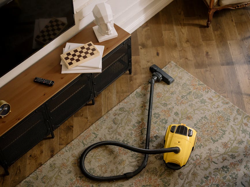 A person holding a yellow portable carpet cleaning or sanitisation device, viewed from behind and slightly above, in a room with a patterned, beige, and blue rug. The device has a large black vent on the front, and the person's hand is adjusting a control on top. The room appears well-lit with soft natural or artificial lighting, and the cleaning equipment is positioned on the floor, suggesting a deep or surface cleaning process in a residential space. The environment is clean, with no visible dust or dirt, and the focus is on the cleaning tool, which may be associated with professional domestic cleaning services by Hounslow Carpet Cleaning, as indicated by the context.