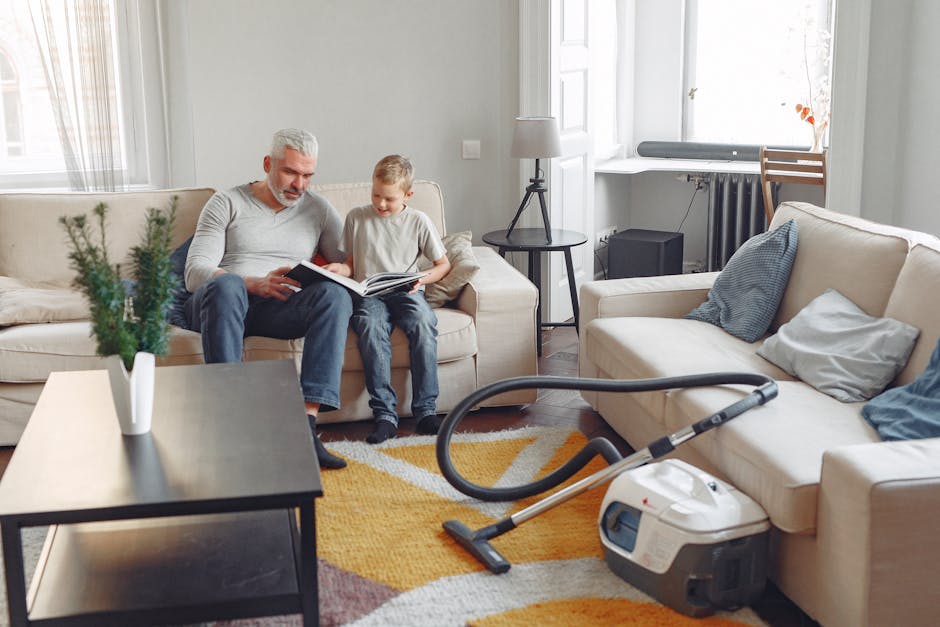 An interior view of a living room featuring a beige fabric sofa with blue and white cushions, a black coffee table with a potted plant, and a vacuum cleaner with a hose on a multicolored rug. The room is illuminated by natural light from large windows with white frames, and has light-colored walls, wooden flooring, a side table with a lamp, and a radiator under the window. The scene depicts a domestic cleaning process, with Hounslow Carpet Cleaning providing surface cleaning and sanitisation services to maintain a hygienic environment.