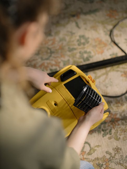 Close-up of a person operating a yellow portable fan or air mover, used for surface drying during deep cleaning or sanitisation. The individual’s hands are visible, with one hand pressing buttons on the device, which has a black vent grille on the front. The fan is positioned on a traditional patterned carpet or rug with floral motifs, partially visible beneath the device. Soft, ambient lighting illuminates the scene, emphasizing the clean, well-maintained appearance of the area. The setting suggests a professional domestic cleaning or surface preparation, and the image is associated with Hounslow Carpet Cleaning, showcasing their surface cleaning and deep cleaning services.