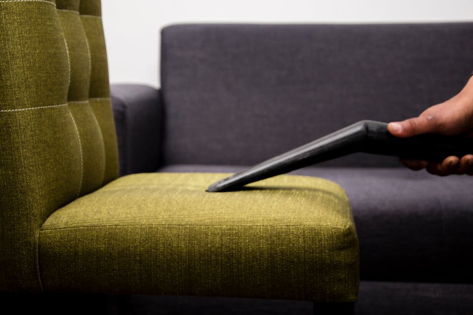 A close-up view of a beige fabric sofa in a living room, featuring tufted cushions with button details and smooth armrests, set against a plain white wall. The sofa has a clean, well-maintained appear