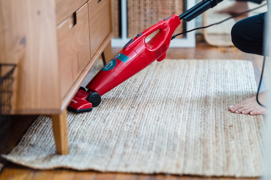 A professional technician wearing black gloves is using a spray cleaning tool connected to a hose to perform deep cleaning and sanitisation on a grey carpeted floor within an indoor space. The area fe