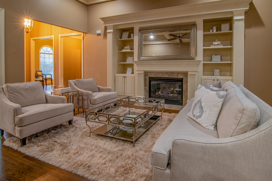 A spacious living room with beige carpet flooring, appearing clean and well-maintained, illuminated by natural light coming through five large, white-framed bay windows adorned with pink and white str