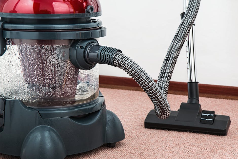 A person kneeling on a patterned carpet in a residential setting, using a yellow portable vacuum cleaner with a black hose attachment. The individual is wearing a beige jacket and jeans, and appears t